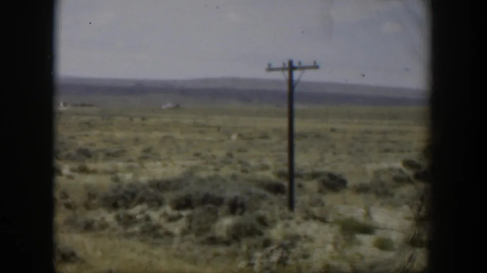 1951 Wyoming landscape frame showing a utility pole, sparse roadside brush, open field, and low mountains in soft 8mm color.