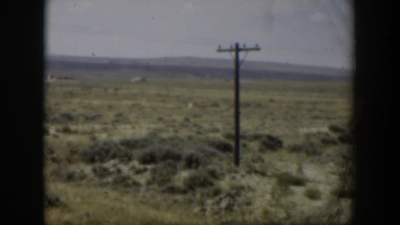 1951 Wyoming 8mm view of a utility pole rising from dry open scrubland, with soft distant hills and dark frame edges.