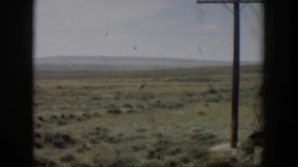 Soft 8mm frame of Wyoming prairie with a faint antelope, scrub vegetation, horizon line, and telephone pole at right.