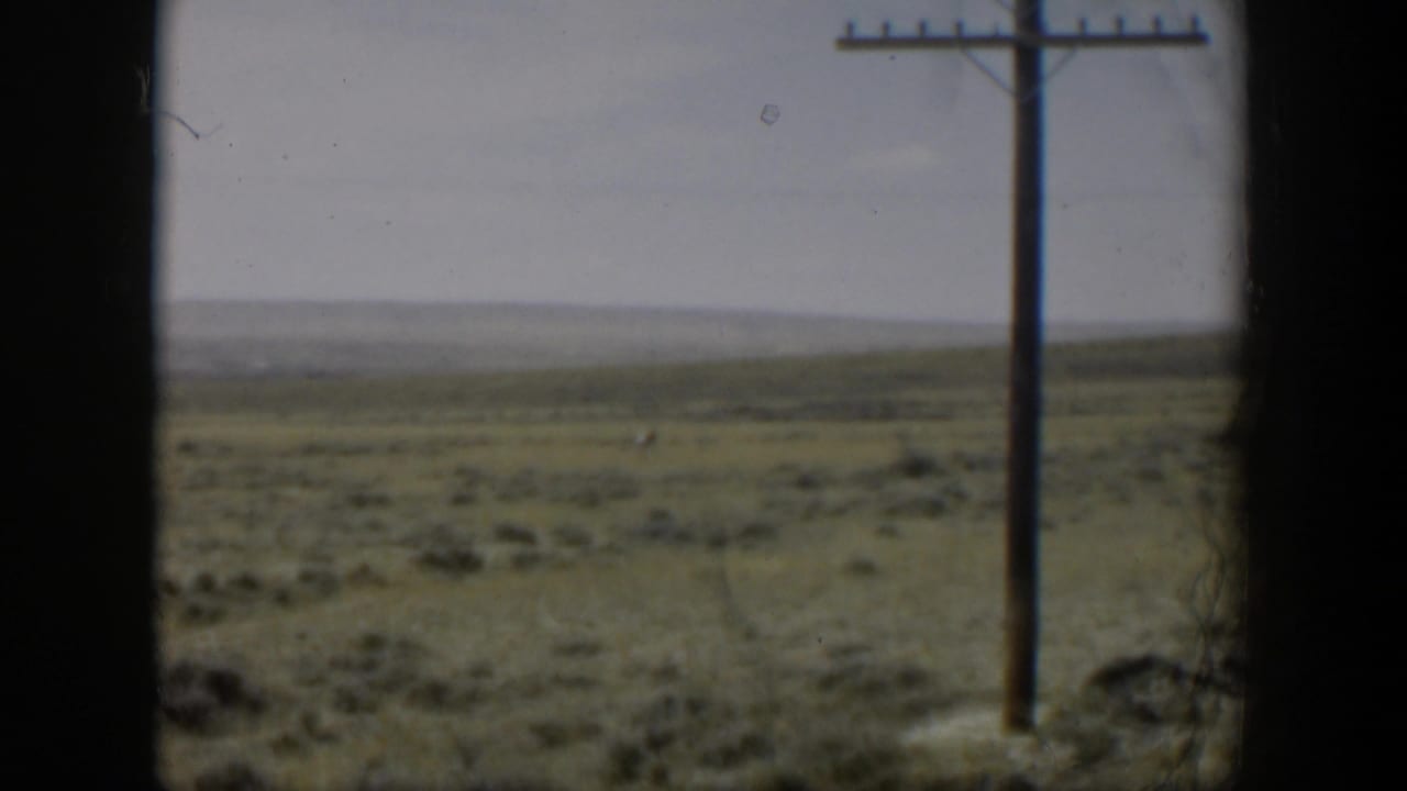 Open plain and utility pole viewed through a dark window frame, with distant land under a gray sky.