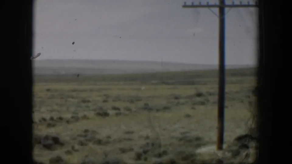 1951 Wyoming 8mm still showing muted grassland through a dark window frame, with a pole and distant plain beyond.
