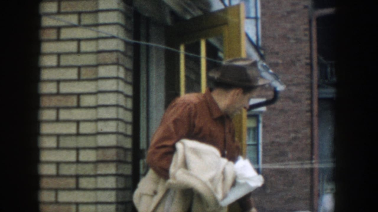 In 1957 Michigan home-movie footage, a man in a brown fedora carries a light coat while exiting a brick building doorway.