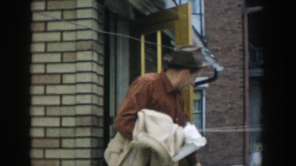 In 1957 Michigan home-movie footage, a man in a brown fedora carries a light coat while exiting a brick building doorway.