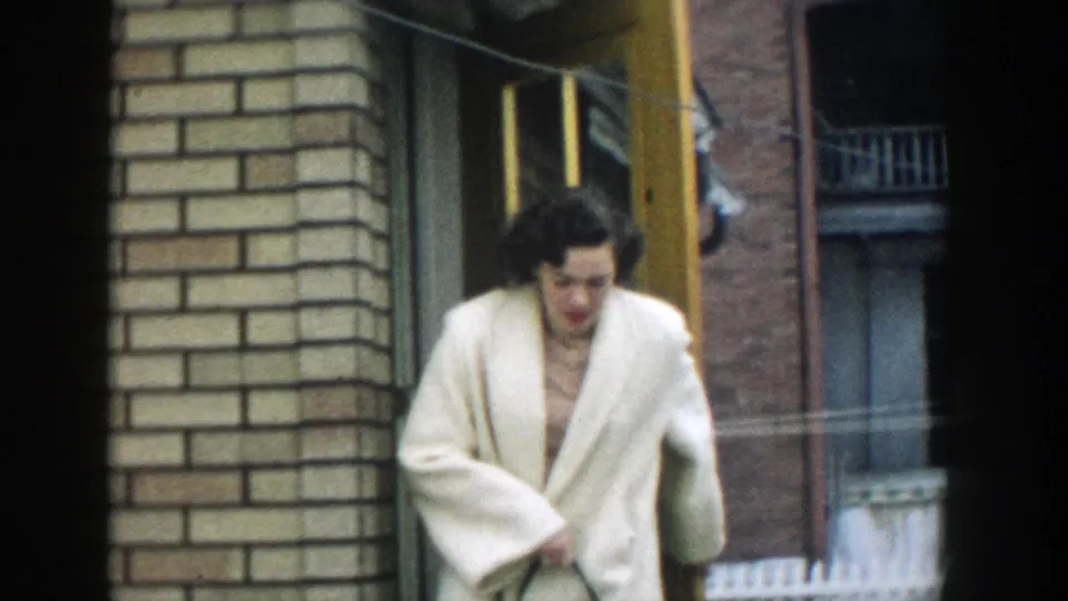 A woman with dark hair steps out from the yellow-framed doorway in 1957 Michigan footage, wearing a white winter coat.
