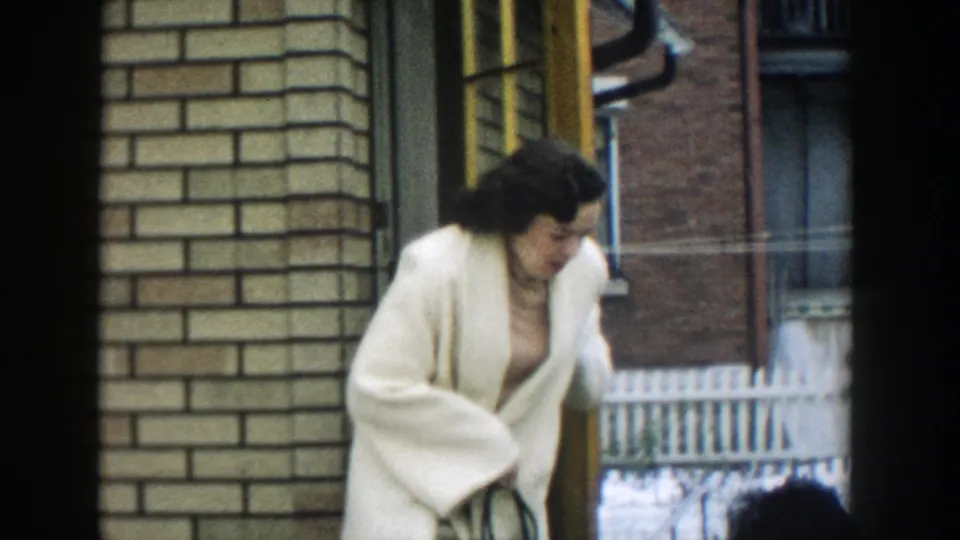 The woman continues away from the doorway in a white coat, with brick walls, windows, and railings visible in the urban residential setting.