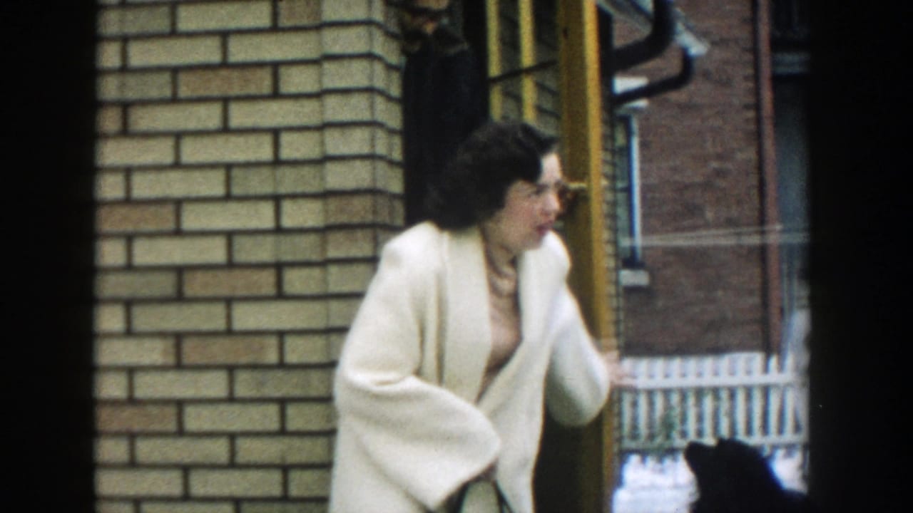 Woman in a cream coat stands near a brick house doorway with a small dog partly visible.