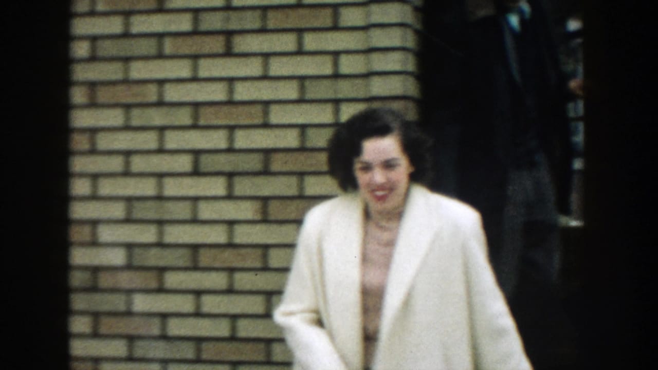 A woman in a cream coat walks out from a brick house in the 1957 Michigan home movie, smiling as she moves past the camera.