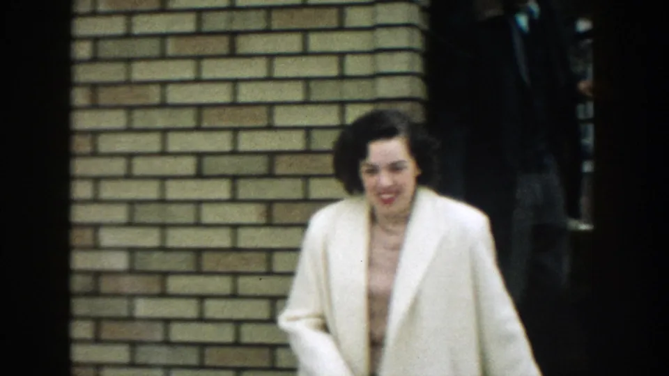A woman in a cream coat walks out from a brick house in the 1957 Michigan home movie, smiling as she moves past the camera.