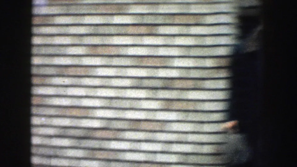 A partially blurred person moves past a light brick wall, with handheld framing and soft 8mm motion obscuring details.
