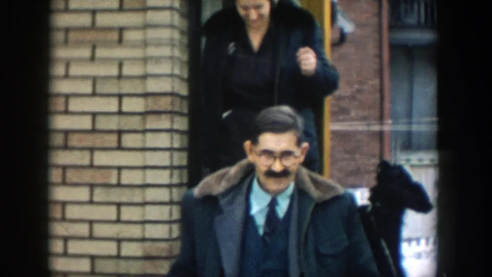 A man wearing glasses and a dark coat walks from the brick house as a woman follows behind and a small dark dog appears at the doorway.