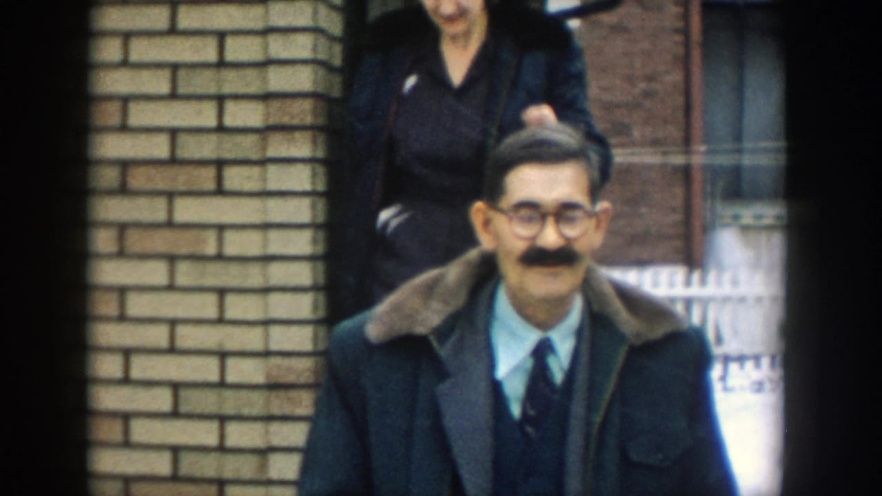 In supported 1957 Michigan home-movie footage, a man in glasses, tie, and dark coat with a fur collar stands near a brick house entrance as another adult is partly visible behind him.