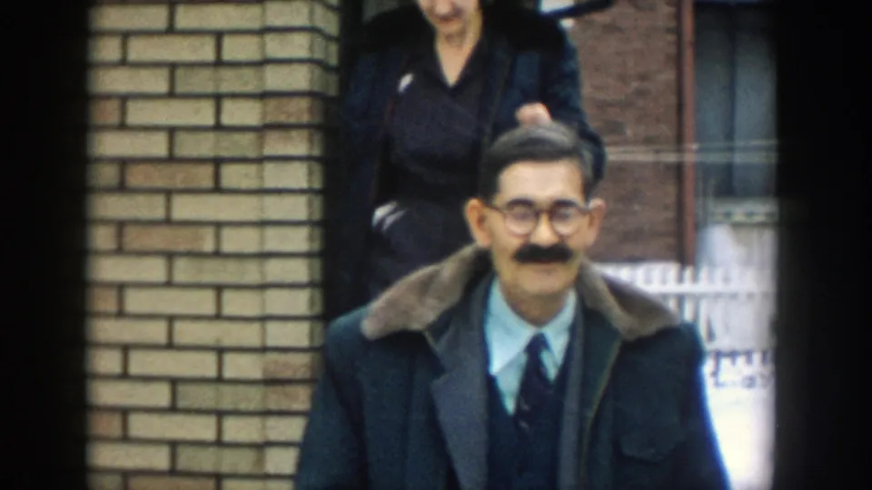 In supported 1957 Michigan home-movie footage, a man in glasses, tie, and dark coat with a fur collar stands near a brick house entrance as another adult is partly visible behind him.
