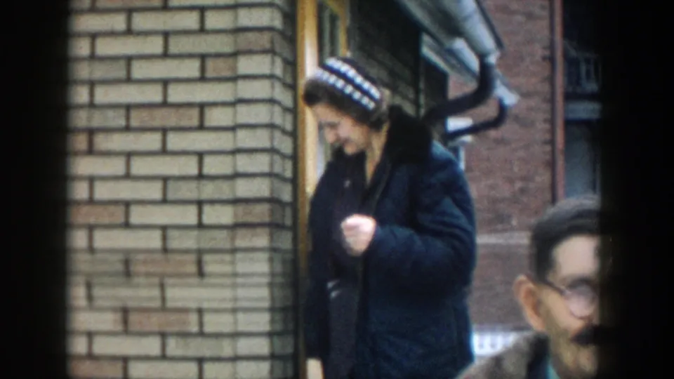 A woman wearing a patterned winter hat and dark coat walks beside a brick house entrance in the 1957 Michigan residential exterior scene, with a man's face partly cropped at the edge.