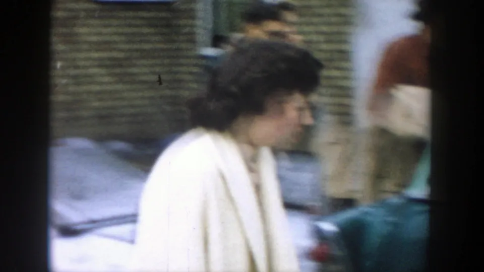 A woman in a light-colored coat moves past a parked car while background figures blur in the handheld 8mm residential scene.