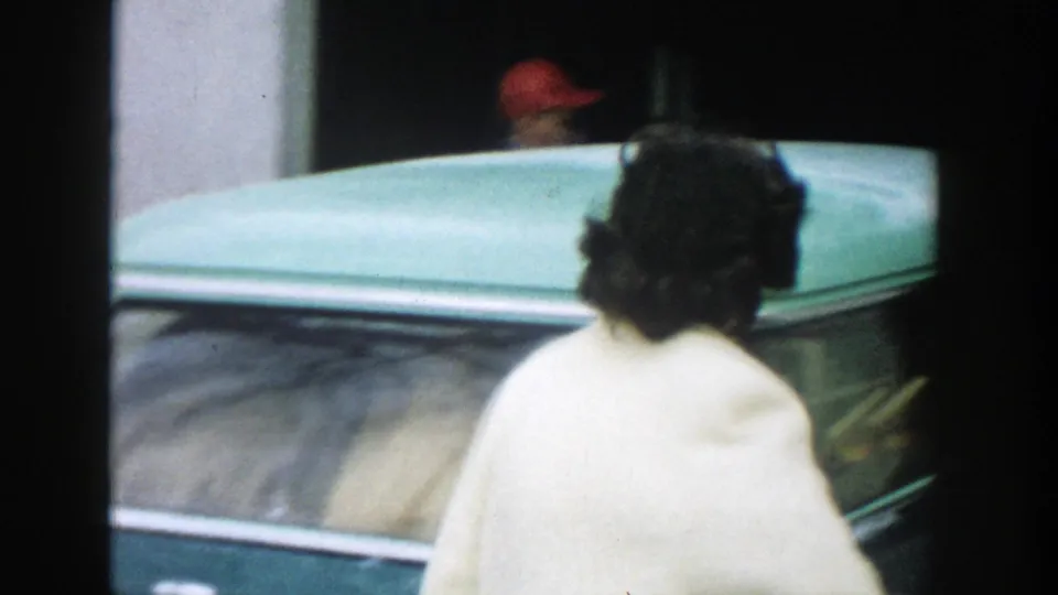 A woman seen from behind in a light coat stands or walks close to a light-colored period car in the Michigan 1957 home-movie exterior.