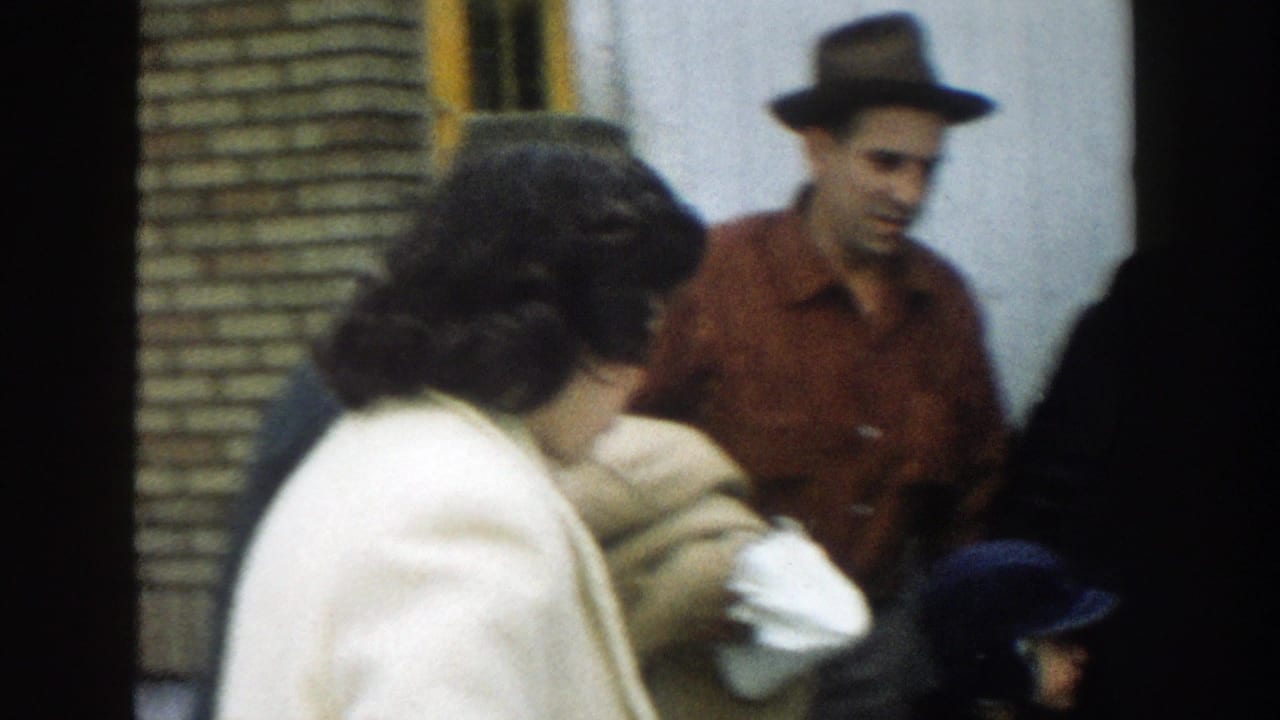 Adults gather outside a building as a man in a brimmed hat stands near women in coats.