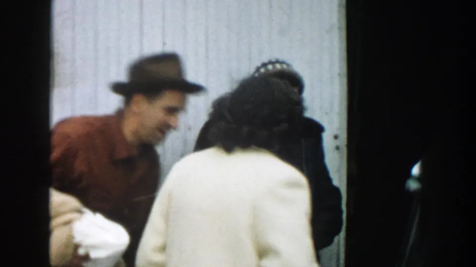 1957 Michigan home-movie frame showing a man in a brown shirt and hat leaning toward women in coats outside a building.