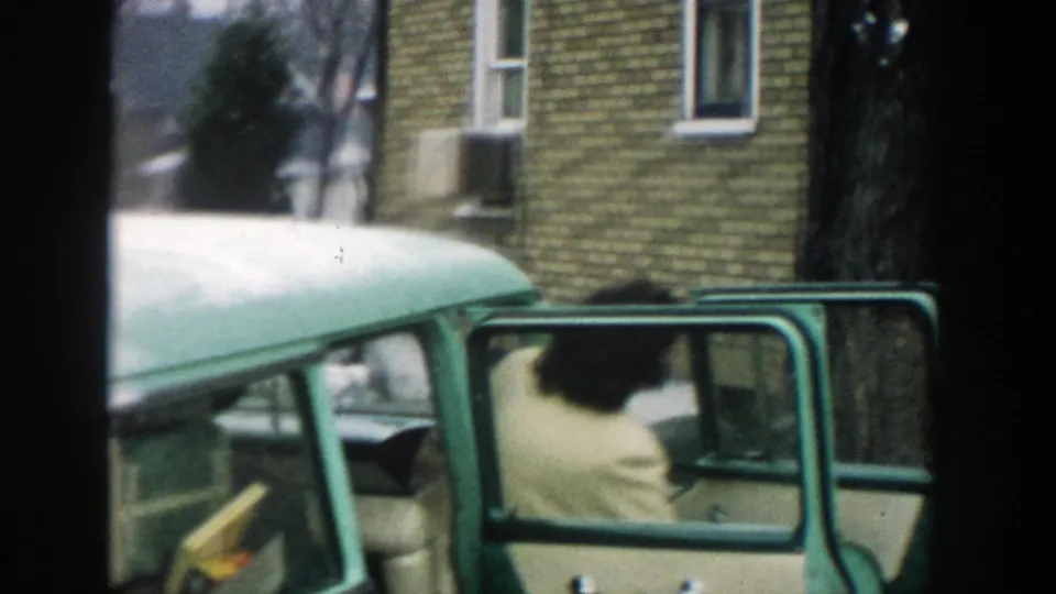 An adult in a light-colored coat appears at the open rear door of the green station wagon in this Michigan home-movie frame.