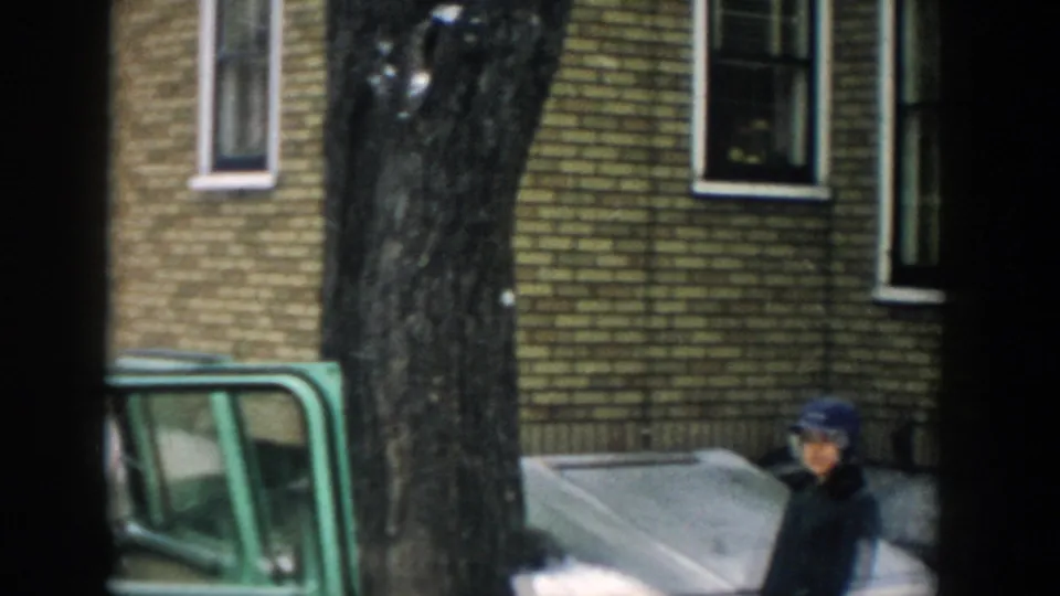 A green station wagon sits by a brick house and large tree, with a child-sized figure visible near the lower right of the frame.