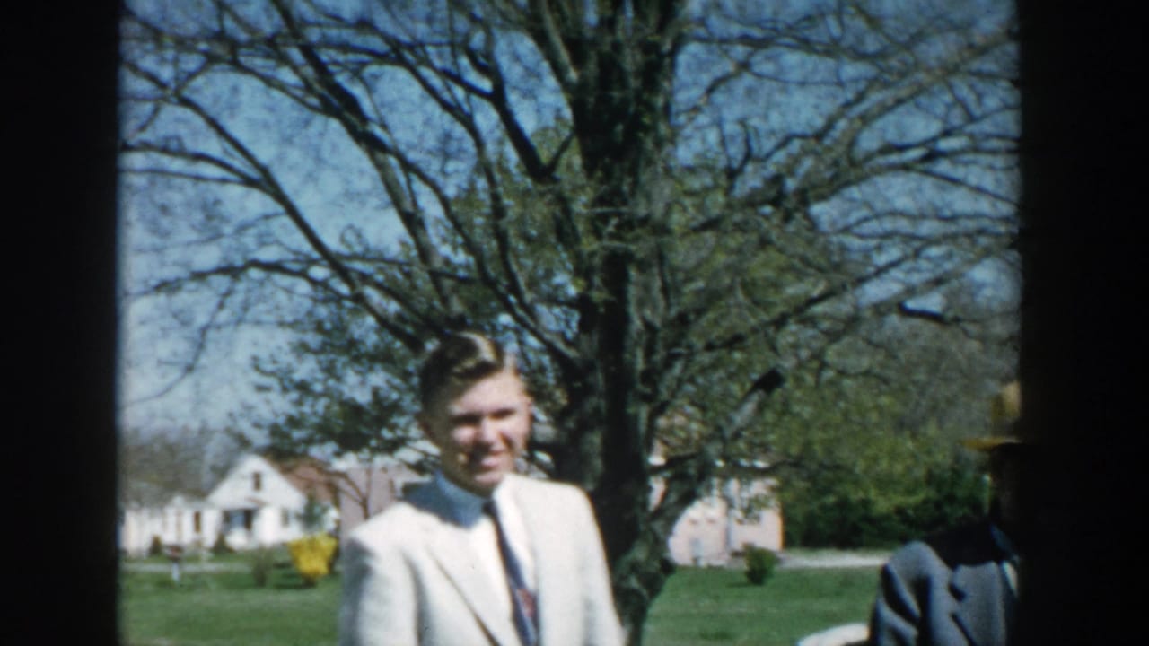 Young man in a light suit smiles outdoors in a suburban yard.