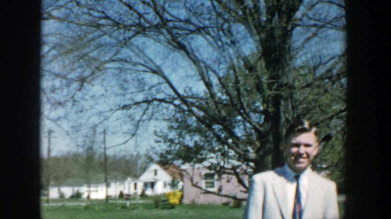 The man in the light suit remains near the right edge, with Michigan lawns, houses, trees, and utility poles behind him.