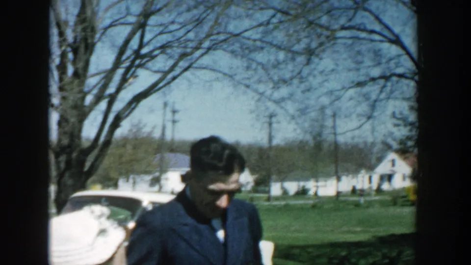 A man in a dark jacket looks down in a Michigan suburban yard, with a woman in a white hat partly visible in front of him.