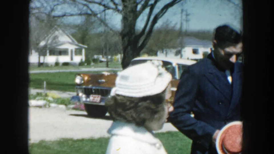 A woman in a white hat and a man in a dark jacket stand near a brown-and-white car in the 1957 Michigan suburban scene.