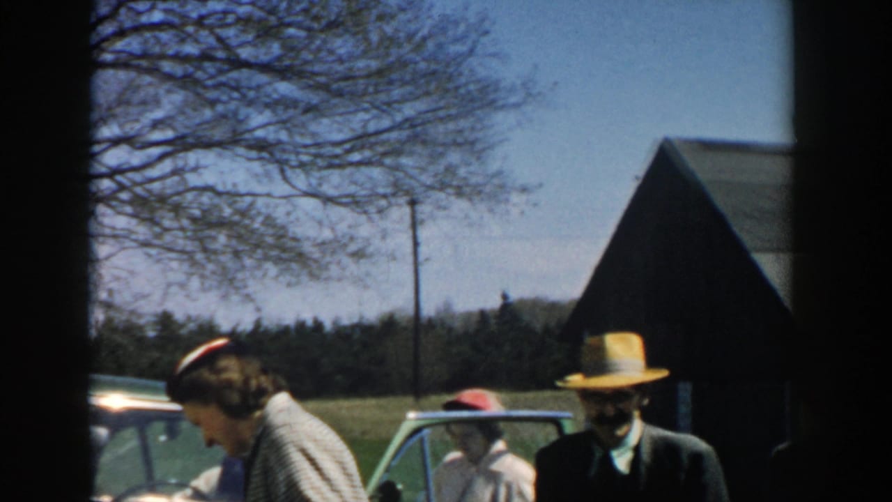 1957 Michigan home-movie frame showing adults in dress clothes near a green car, with a barn-like building, open field, and leafless trees behind them.