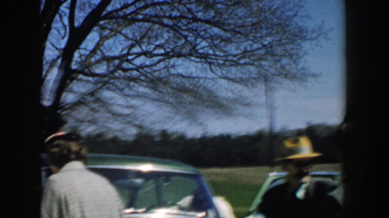 1957 Michigan frame of adults near parked cars in a rural field, including a man in a brimmed hat and a woman seen from behind.