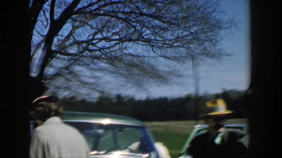 1957 Michigan frame of adults near parked cars in a rural field, including a man in a brimmed hat and a woman seen from behind.