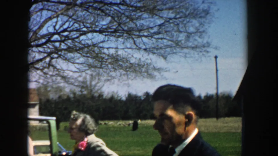 1957 Michigan home-movie frame with a suited man in profile and an older woman near an open car door, set against grass, trees, and sky.