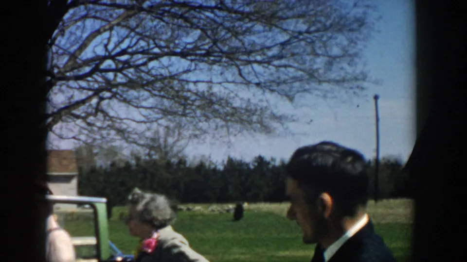 1957 Michigan frame showing a suited man in profile near a car, with an older woman, open field, utility pole, and leafless branches in view.