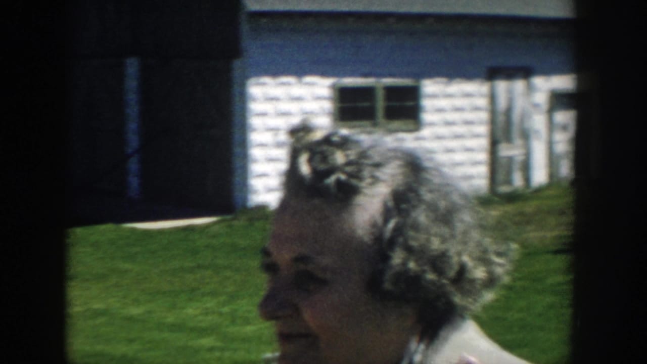 Adult woman with styled gray hair stands in profile near a white outbuilding on a sunny Michigan lawn in 1957 home-movie footage.