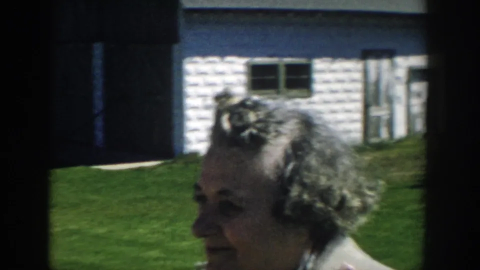 Adult woman with styled gray hair stands in profile near a white outbuilding on a sunny Michigan lawn in 1957 home-movie footage.