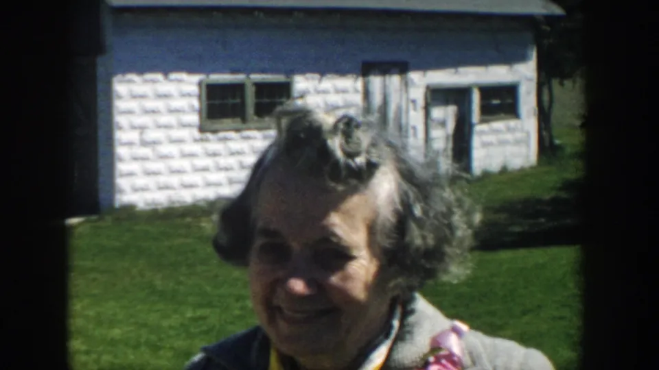 Smiling adult woman faces the camera outdoors, with a textured white shed and green lawn behind her in Michigan 1957 footage.