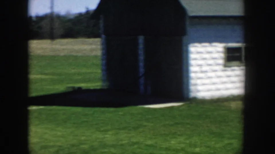 White outbuilding, open lawn, and distant tree line appear in a sunny Michigan exterior from the 1957 home-movie clip.