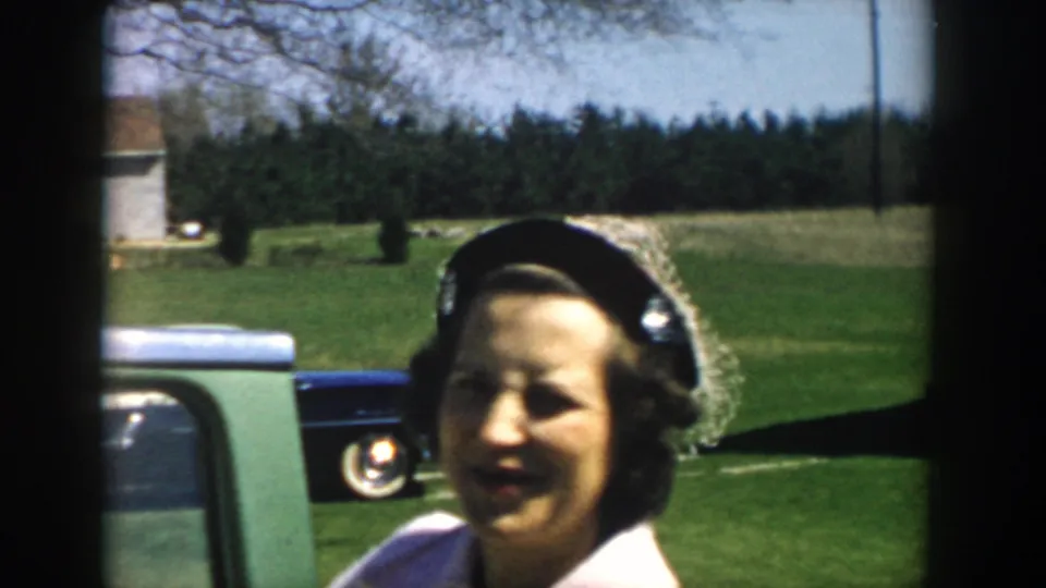Adult woman smiles beside or near a blue-green car, with lawn, trees, and a white building in Michigan 1957 home-movie footage.