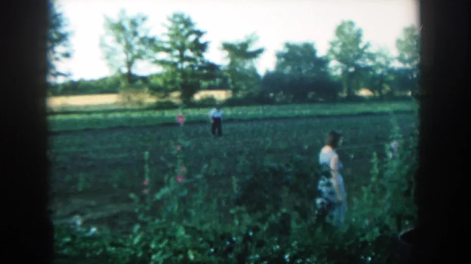 The woman stands beside dense garden plants on a rural Michigan farm, with a man in a white shirt farther back in the field.