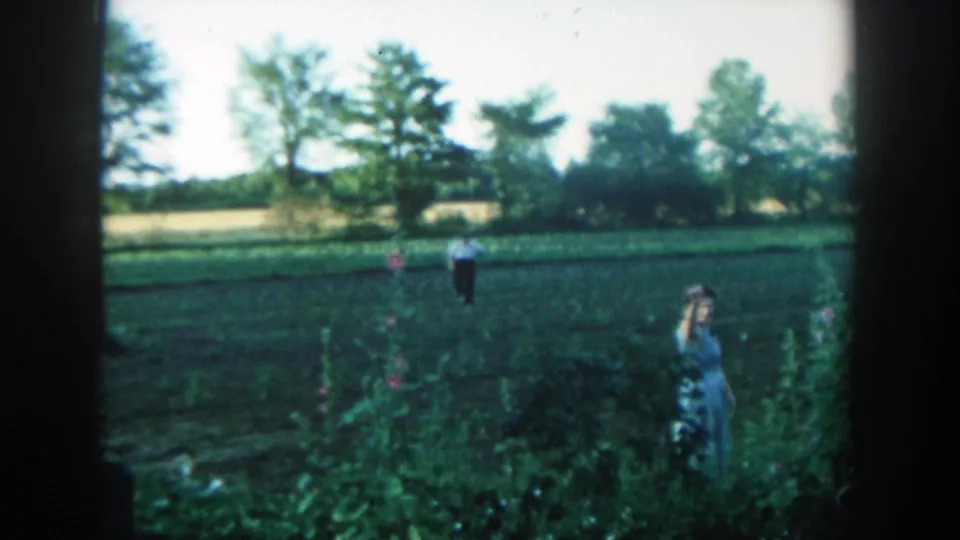 On a 1957 Michigan farm, the woman appears to wave near the garden edge as the man remains in the cultivated field behind her.