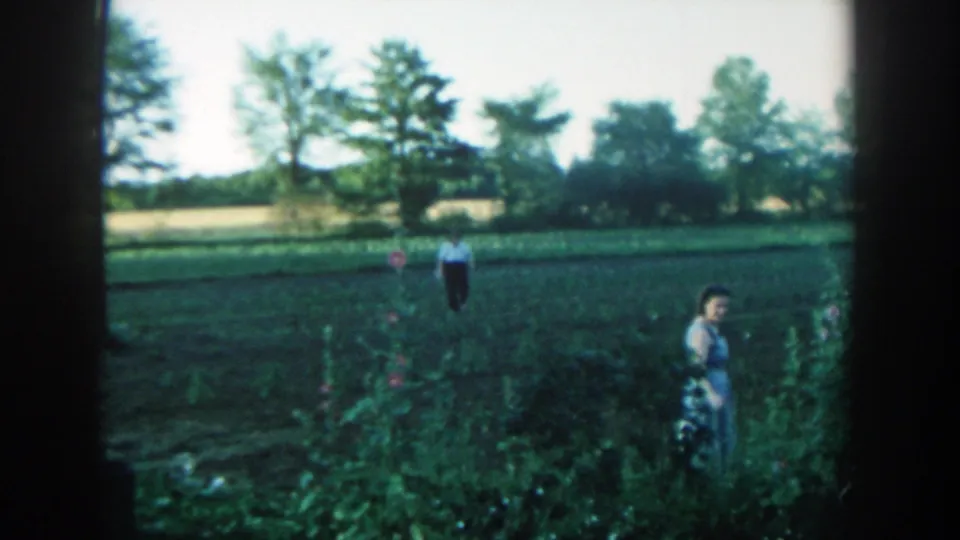 The woman turns toward the camera near flowers and tall greenery, with the man centered farther back in the Michigan farm field.