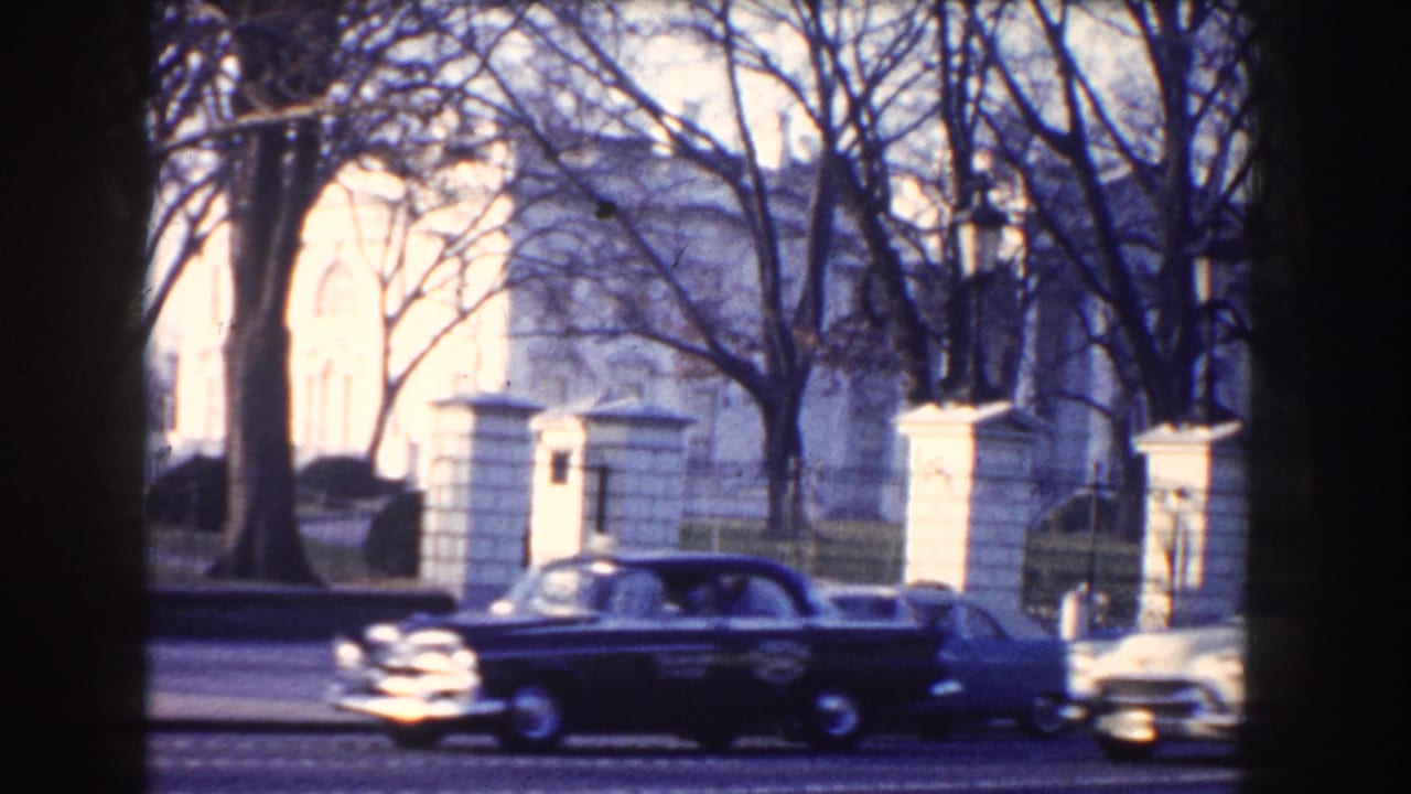 Dark sedan passing in front of the White House behind bare trees.