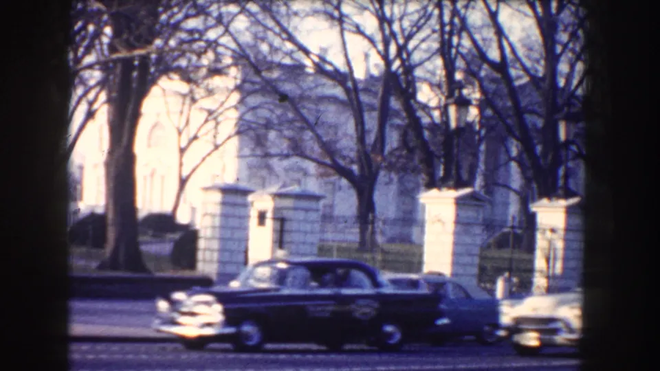 1960 Washington, DC home-movie frame of a dark mid-century sedan moving past the White House through bare winter trees.