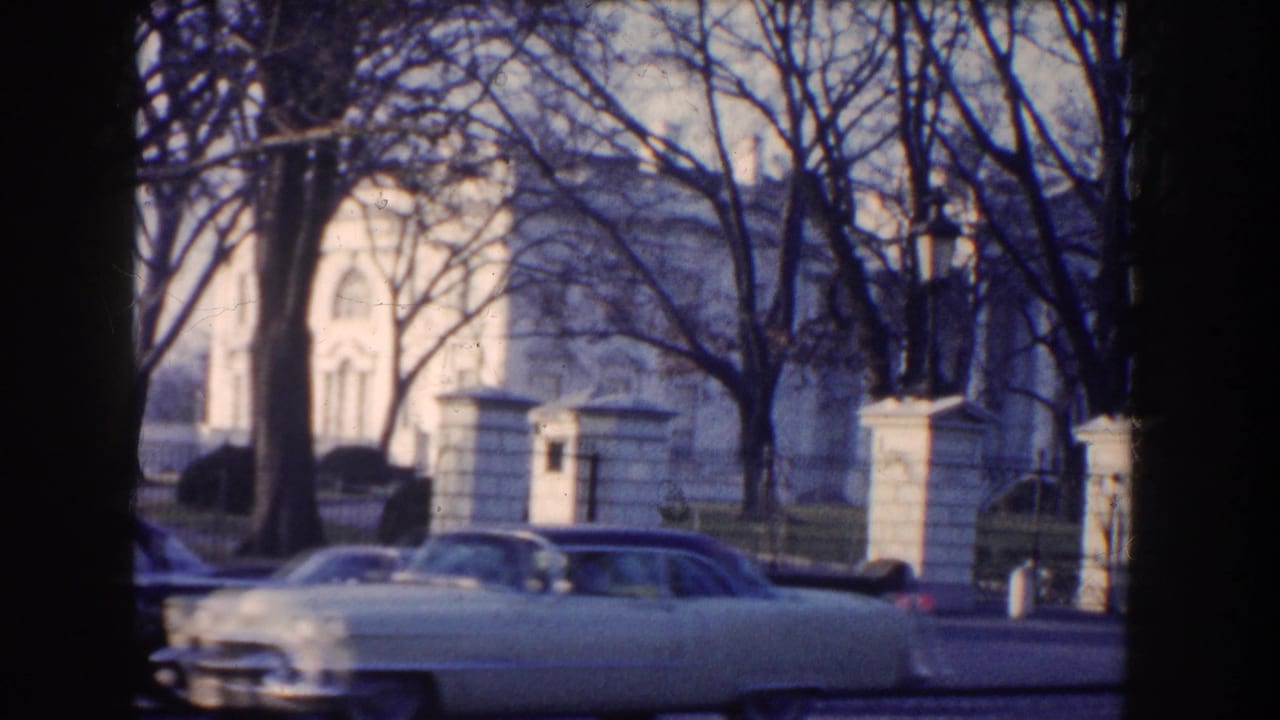 A light-colored 1950s-style sedan passes the White House grounds in 1960 Washington, DC, partly obscured by tree trunks.