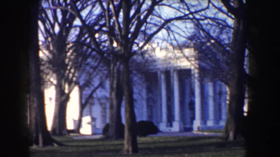 Distant White House view framed by bare trees and lawn in Washington, D.C., on blue-toned 8mm footage from the 1960 clip.