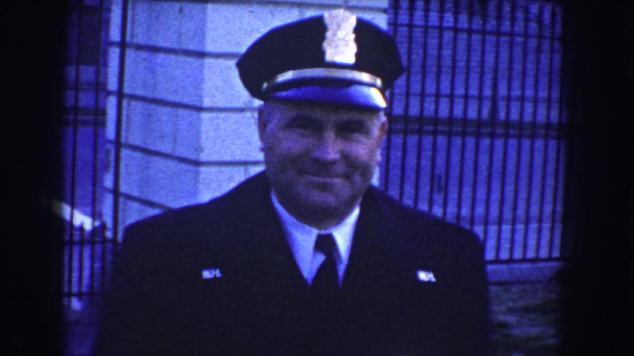 Smiling police officer in dark uniform and peaked cap stands near iron fencing and stone building details in Washington, D.C., circa 1960.