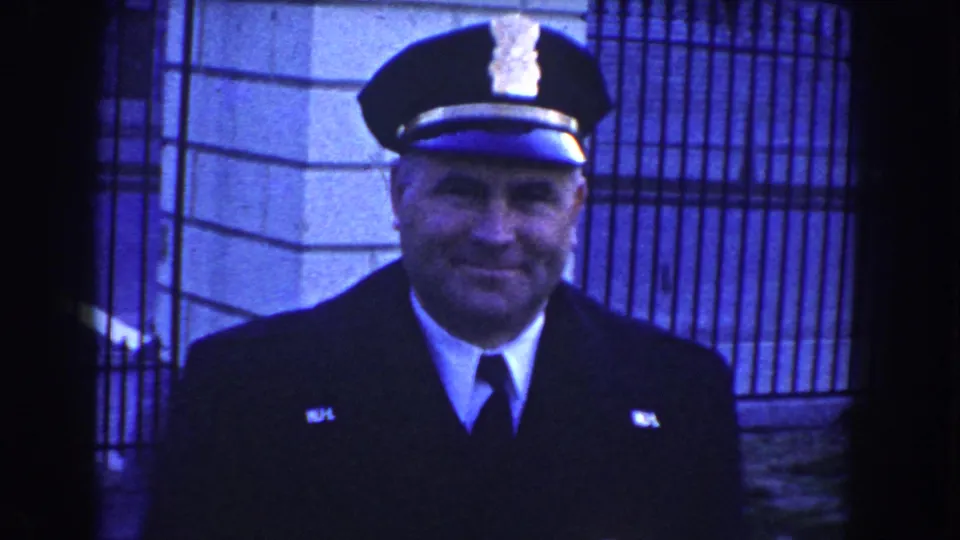 Smiling police officer in dark uniform and peaked cap stands near iron fencing and stone building details in Washington, D.C., circa 1960.