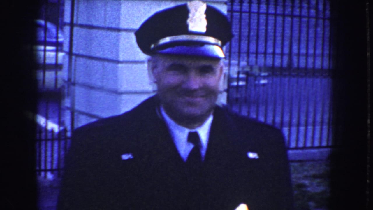 Uniformed police officer smiles toward the camera in front of iron fencing and a stone exterior, likely near the White House grounds in Washington, D.C.