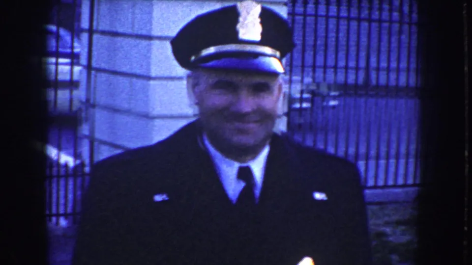 Uniformed police officer smiles toward the camera in front of iron fencing and a stone exterior, likely near the White House grounds in Washington, D.C.