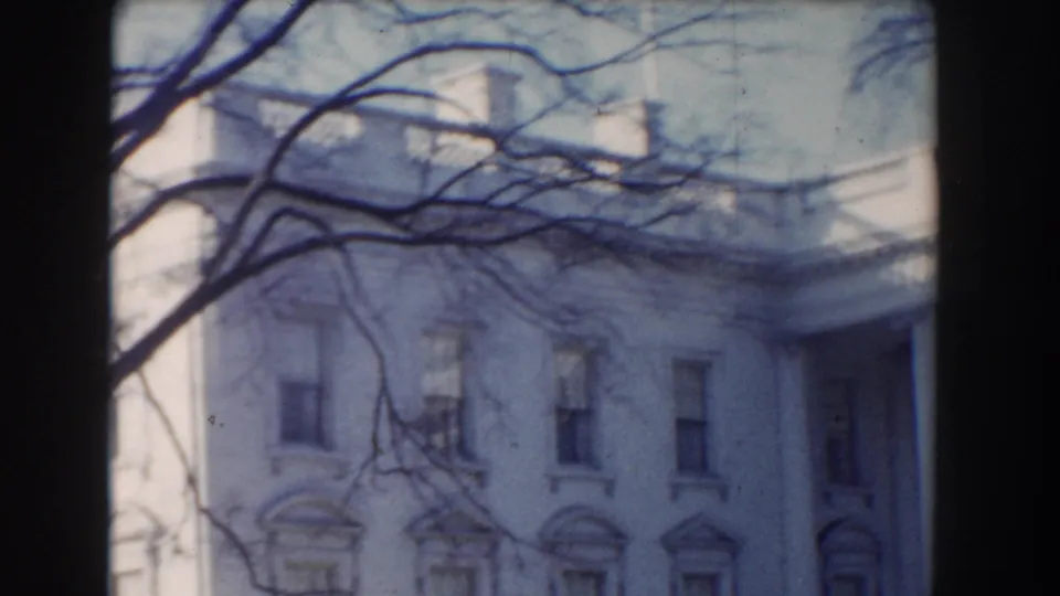 Soft 8mm frame of the White House exterior, with upper facade details and leafless branches crossing the view.