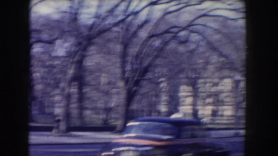 A 1960-era car moves past a Washington DC sidewalk with leafless trees and a pale building behind fencing.